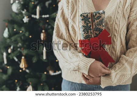 Hands in cozy sweater holding stylish christmas gift in festive wrapping paper with bow on background of christmas tree lights. Happy holidays! Atmospheric christmas time