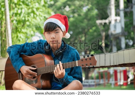 asian cute boy wears santa hat sitting and playing ukulele or acoustic guitar in puplic park, concept for lifestyle of children in special holidays, christmas day and new year events around the world.
