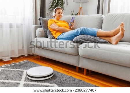 A woman relaxes on a comfortable sofa, while a robot vacuum cleans the room Royalty-Free Stock Photo #2380277407