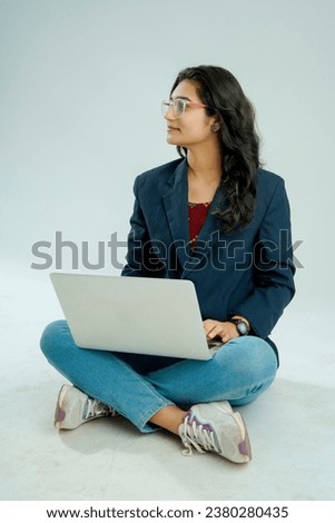 indian girl seated on a white background balances a laptop on her lap, gesturing while she speaks, displaying both multitasking skill and expressive communication.