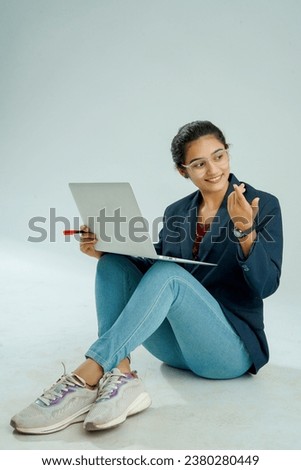 indian girl seated on a white background balances a laptop on her lap, gesturing while she speaks, displaying both multitasking skill and expressive communication.