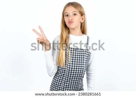 caucasian kid girl wearing dress makes peace gesture keeps lips folded shows v sign. Body language concept