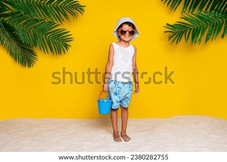 Full length photo of funky cool little boy dressed white shirt dark eyewear playing sand isolated yellow color background