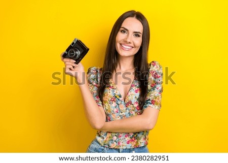 Photo of satisfied cheerful girl with long hairdo dressed flower print blouse hand holding camera isolated on yellow color background