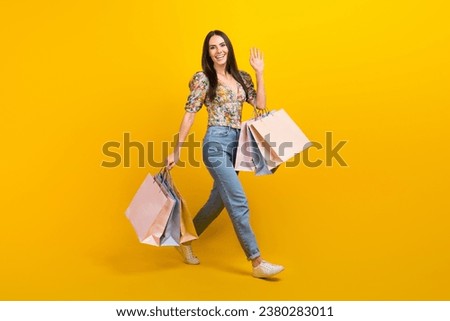 Full length photo of pretty girl dressed flower print blouse hold new clothes say hello waving arm isolated on yellow color background