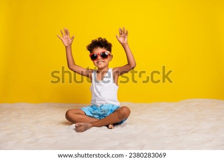 Full length photo of sweet cheerful small boy wear white shirt dark eyewear siting beach rising arms empty space isolated yellow color background Royalty-Free Stock Photo #2380283069