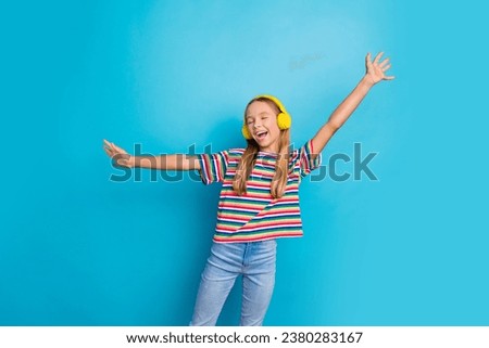 Portrait of carefree overjoyed teen girl wear colorful t-shirt in headphones raisng arms up have fun isolated on blue color background Royalty-Free Stock Photo #2380283167