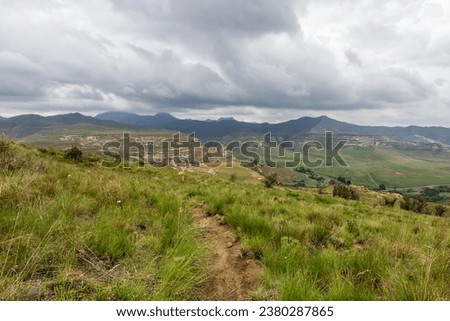 A small hiking trail high up in the Drakensberg Mountains of South Africa, with storm clouds gathering over the distant blue peaks in the background Royalty-Free Stock Photo #2380287865