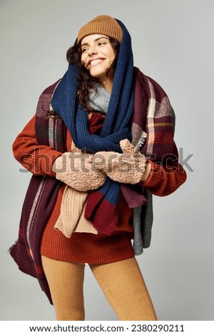 winter fashion, joyous model in layered clothing, warm hat and scarfs posing on grey backdrop Royalty-Free Stock Photo #2380290211