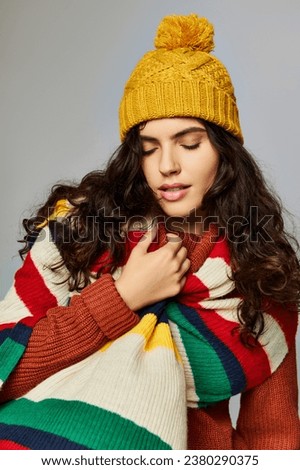 brunette woman in winter bobble hat and sweater with stripped scarf posing on grey background