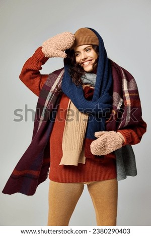 winter style, happy young woman in layered clothing, warm hat and scarfs posing on grey backdrop