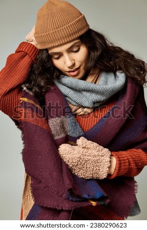 winter fashion, curly brunette woman in layered clothes with warm scarf and mittens on grey backdrop Royalty-Free Stock Photo #2380290623