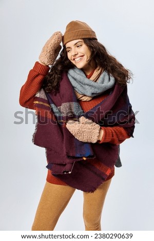winter fashion, happy curly woman in layered clothes with warm scarf and mittens on grey backdrop