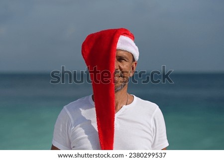 Portrait of a tanned man in a white T-shirt and Santa Claus hat with a view of the blue ocean. Santa Claus hat covers half of the face. Christmas trip to the hot countries of Santa Claus