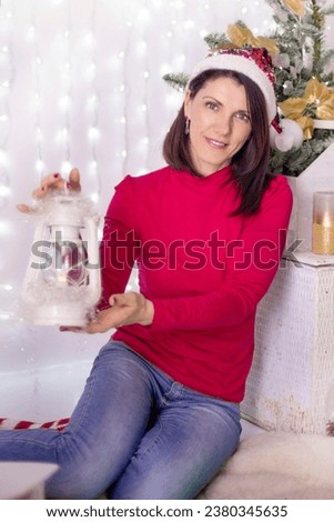girl in a Christmas hat holds fairy lights, smiles, poses among beautiful New Year's decor, holiday lights, Decorated Christmas tree, balls.
