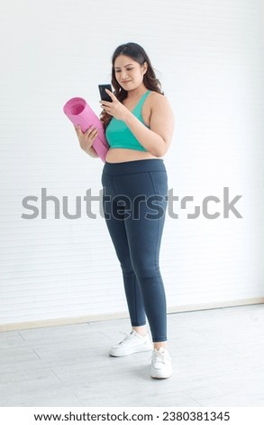 Asia young chubby fat healthy oversized overweight plus size female sportswoman in sportswear sports bra legging sneakers standing holding yoga pilates mat looking at water bottle in home fitness gym. Royalty-Free Stock Photo #2380381345