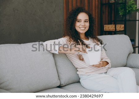 Side view young woman in casual clothes sit on grey sofa couch eat Chinese food cuisine in takeaway carton container box stay at home hotel flat rest relax spend free spare time in living room indoor