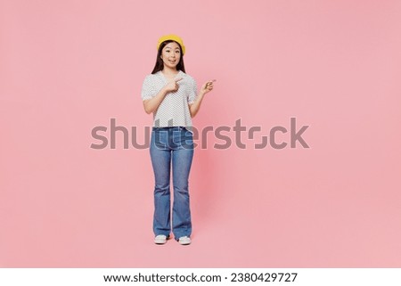 Full body young woman of Asian ethnicity wear white polka dot t-shirt yellow beret point index finger aside on workspace area mock up isolated on plain pastel pink background. People lifestyle concept