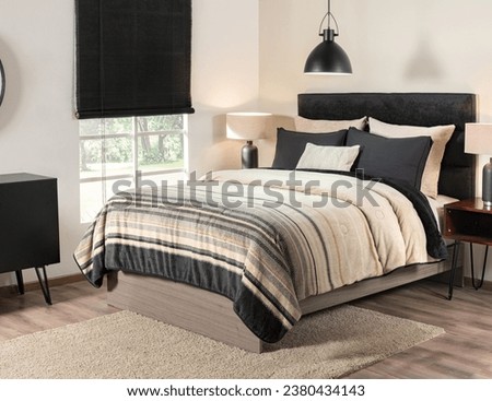 A Nordic-style bedroom with a black upholstered headboard, pillows, a striped quilt in beige and black tones, side tables, a window with a garden view, pendant light, all atop a beige area rug. Royalty-Free Stock Photo #2380434143