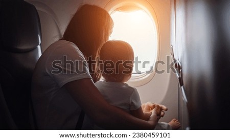 Joyful mother shows view from plane window to toddler girl. Child cradled in arms of mother as family embarks on memorable vacation together, sunlight Royalty-Free Stock Photo #2380459101
