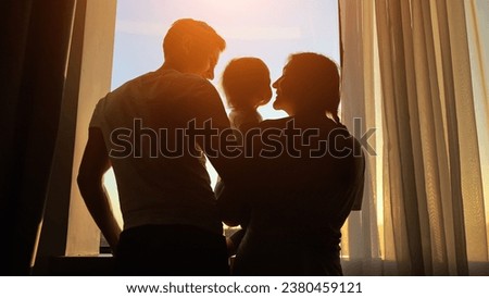 Mother holds toddler in arms near window with husband watching sunset. Family admires view mom leans down to plant kissing child cheek