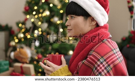 Young chinese woman drinking coffee sitting on sofa by christmas tree at home