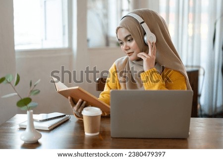 A relaxed and charming Asian-Muslim woman wearing a hijab and headphones is listening to music and reading a book while working remotely at a cafe.