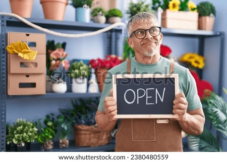 Hispanic man with grey hair working at florist holding open sign smiling looking to the side and staring away thinking. 