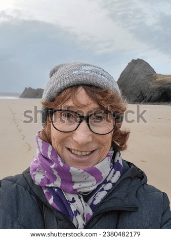 Mature woman walking on Three Cliffs Bay in winter takes a selfie. She is wearing warm winter clothing including a bobble hat and scarf. The three cliffs are behind her. 
