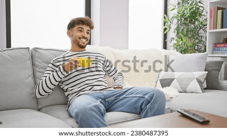 Chilled-out young arab man smiling as he enjoys his morning espresso, comfortably relaxed watching a film on his tv, living room background.