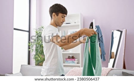 Handsome young hispanic man standing in the comfort of his living room, focused on folding laundry from a towel basket, creating a picture of relaxation amidst household chores.