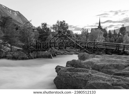 Bridge crossing a  big river flowing through the Lom village. Image taken during summer night. Slow shutter speed long exposure image. Old wooden church on the background. River through the cliffs