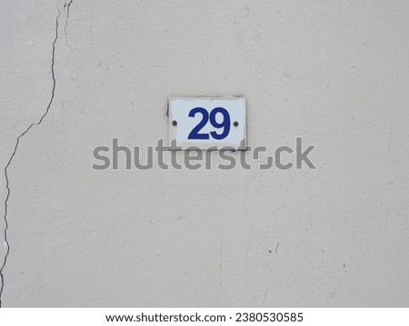      A small square plaque with the numbers twenty-nine attached to a white wall with cracked plaster     