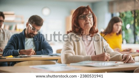 Portrait of Happy Senior Woman Taking a Course in an International Adult Education Center. Old Female Wearing Glasses, Sitting Behind a Desk, Using Textbook and Writing Down Notes in Notebook