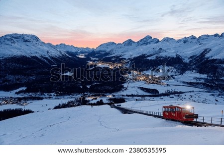 A funicular carriage on the snowy slope toward Muottas Muragl at dusk, overlooking St Motritz, Celerina, Pontresina and lakes in Engadin Valley under alpine mountains, in Samedan, Grisons, Switzerland Royalty-Free Stock Photo #2380535595