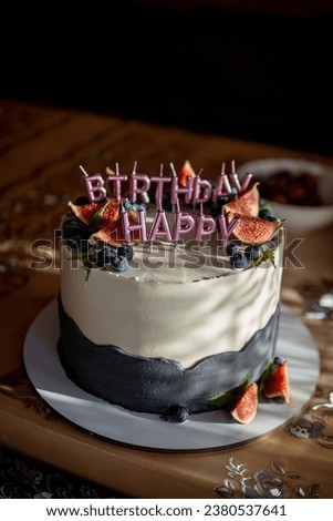 A round cake is on the table. Vertical photo. Candles in the form of letters Happy Birthday. Decorated with berries and fruits, blueberries and dates. White blue color Presents at birthday party.