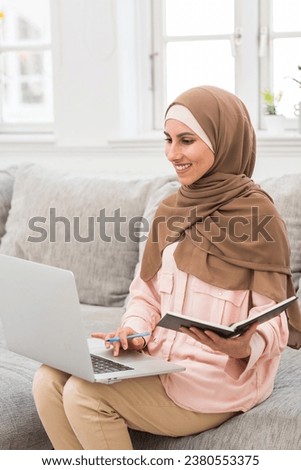 Vertical photo of a cute Arab girl in hijab sitting in a cozy atmosphere in the living room on the sofa, holding a laptop in her hand and taking notes on a webinar, teaching or working at home.