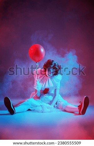 Mysterious old circus. A clown girl in a white circus dress sits on a dark stage, shrouded in haze, and looks intently at the camera, holding a balloon. Halloween.