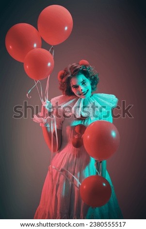 Old circus concept. Portrait of a clown girl in a white circus outfit, who stands on a dark gloomy background in the smoke and smiles, holding red balloons in her hands. Scary Halloween.