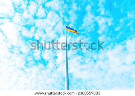 Ukrainian flag, seen from below, as it rises high above the fluffy altocumulus clouds against a serene blue sky.