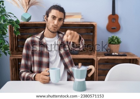 Young hispanic man drinking coffee from french coffee maker pointing with finger to the camera and to you, confident gesture looking serious 