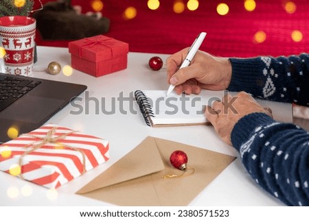 Writing a Christmas Wishing Card. Male adult making greeting cards at home office during Xmas. Man hands in blue sweater holding blank paper and envelope at the Desktop. Coffee Cup, holiday decoration