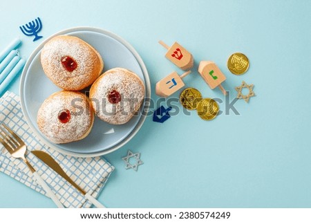 Hanukkah feast presentation. Top view image of Jewish sufganiyot on plate, napkin, cutlery, Star of David, candles, gelt, dreidel set on a pastel blue background with text or ad space