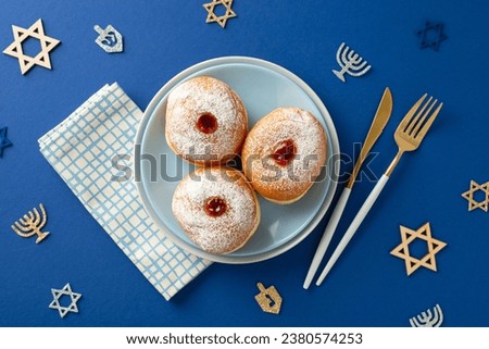 Embrace Hanukkah customs with a top-view image of plate with sufganiyot, napkin, cutlery, Star of David and menorah signs against a blue backdrop, perfect for promotion or advertising