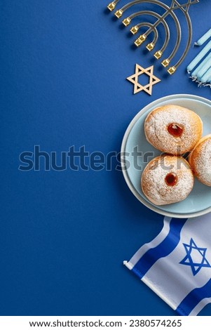 Observe Hanukkah with tasteful table setup. Overhead vertical shot of traditional Jewish dish - sufganiyot on plate, Star of David, Israel flag, menorah on blue backdrop with blank area for your text Royalty-Free Stock Photo #2380574265