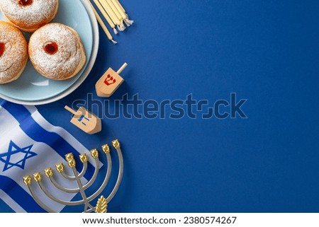Honor the Hanukkah season with a top-view photo featuring sufganiyot, Israeli flag, candles, and dreidel on a blue background, allowing for text or ad inclusion Royalty-Free Stock Photo #2380574267