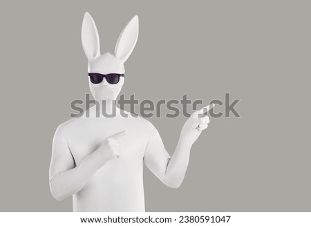 Portrait of faceless unrecognizable person wearing black sunglasses and animal hare mask with long ears in white spandex costume pointing index finger to the side isolated on grey background. Royalty-Free Stock Photo #2380591047