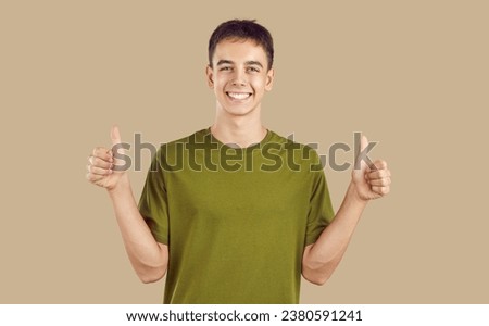 Happy high school or college student giving a thumbs up. Young man or teenage boy in a green tee standing isolated on a beige background, looking at the camera, doing a thumbs up gesture and smiling