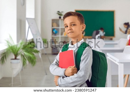 Proud boy first grader with satchel and workbook posing on first day of elementary school getting ready to receive quality assignment standing in classroom with big blackboard. Back to school Royalty-Free Stock Photo #2380591247