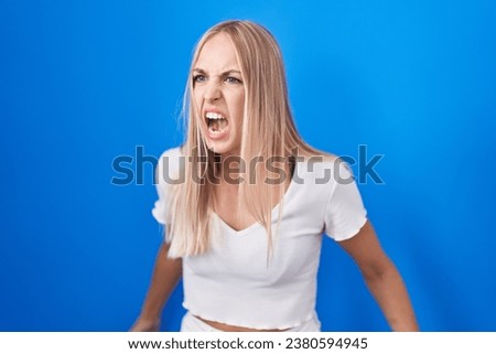 Young caucasian woman standing over blue background angry and mad screaming frustrated and furious, shouting with anger. rage and aggressive concept. 
